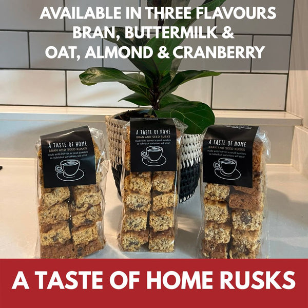 Three bags of A Taste of Home rusks cookies displayed on a kitchen counter with a potted plant and white subway tiles in the background. White text at the top lists available flavors: bran, buttermilk, oat, almond & cranberry.