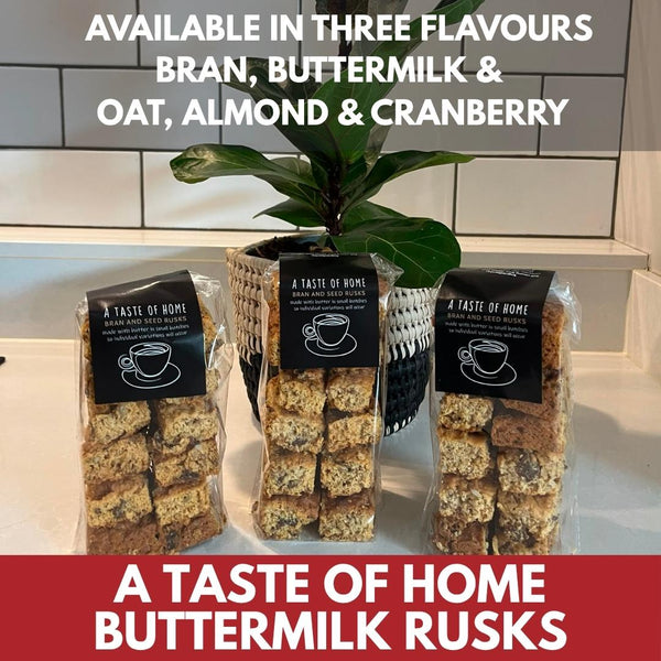 Three clear plastic bags of buttermilk rusks cookies with black labels are displayed on a kitchen counter in front of a tiled wall and a potted plant. Large text overlays read 'AVAILABLE IN THREE BRAN, BUTTERMILK & OAT, ALMOND & CRANBERRY' and 'A TASTE OF HOME BUTTERMILK RUSKS'.