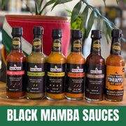 Six bottles of Black Mamba hot sauces lined up on a wooden surface, with a green leafy plant and a red pot in the blurred background. A green banner at the bottom reads 'BLACK MAMBA SAUCES'.