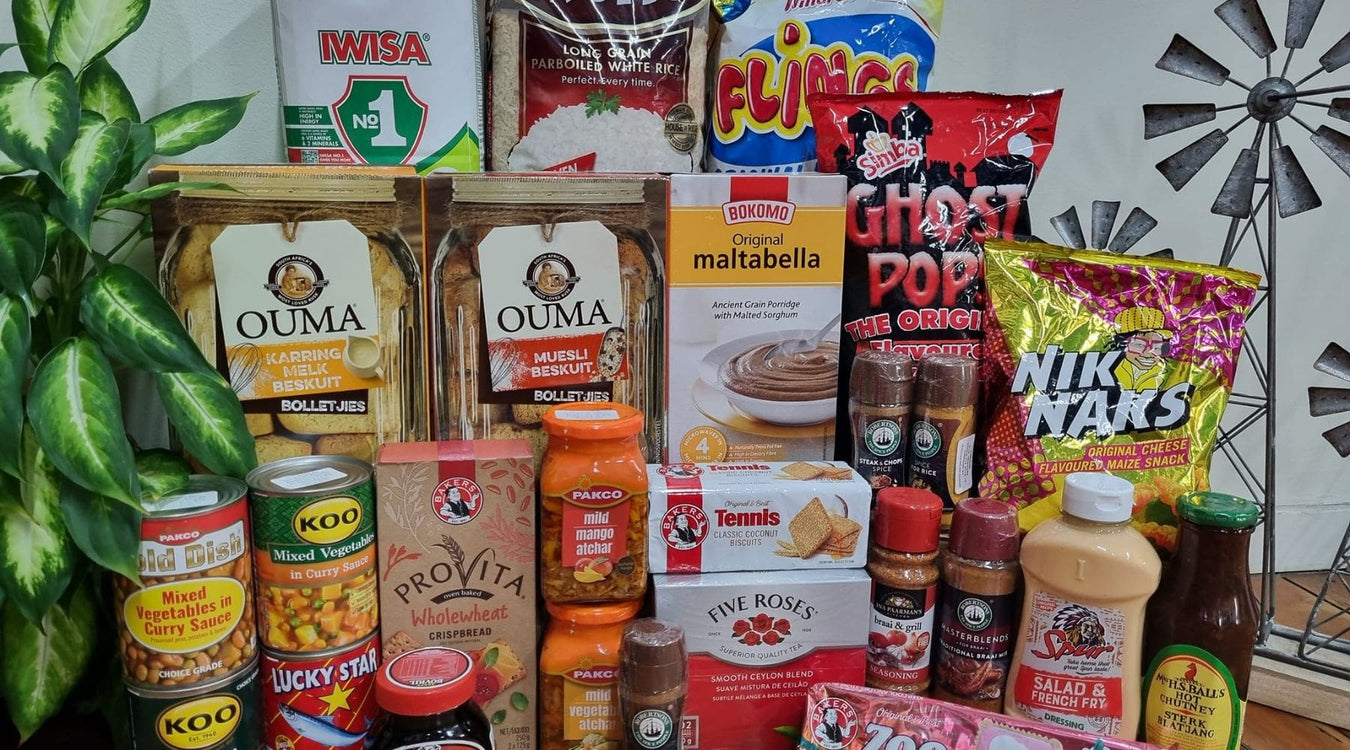 Assorted South African groceries including snacks, sauces, and beverages on a table with a decorative windmill in the background.